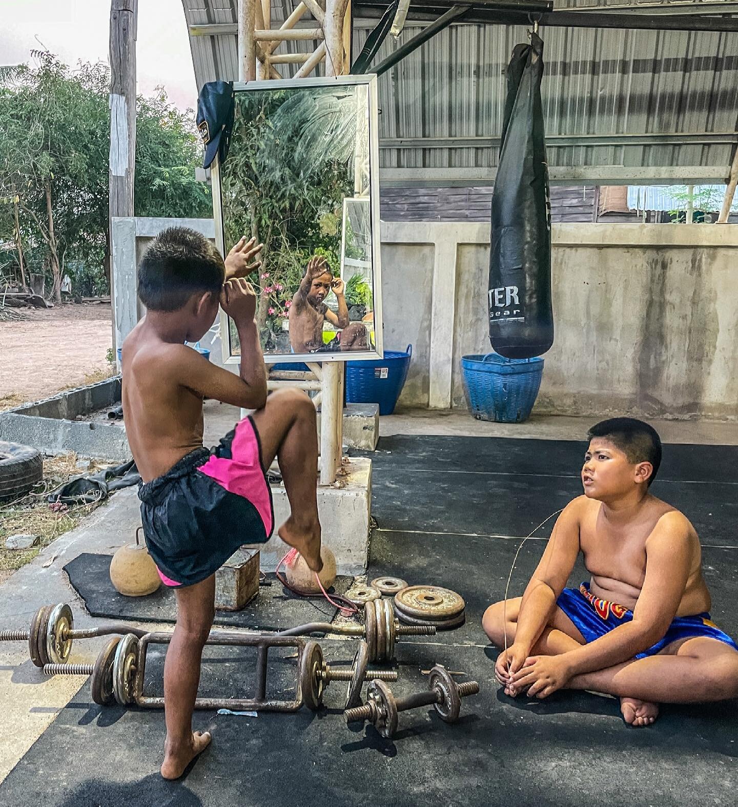 A boy shadowboxing in front of a mirror in the open-air gym while a teammate watches from the mat.