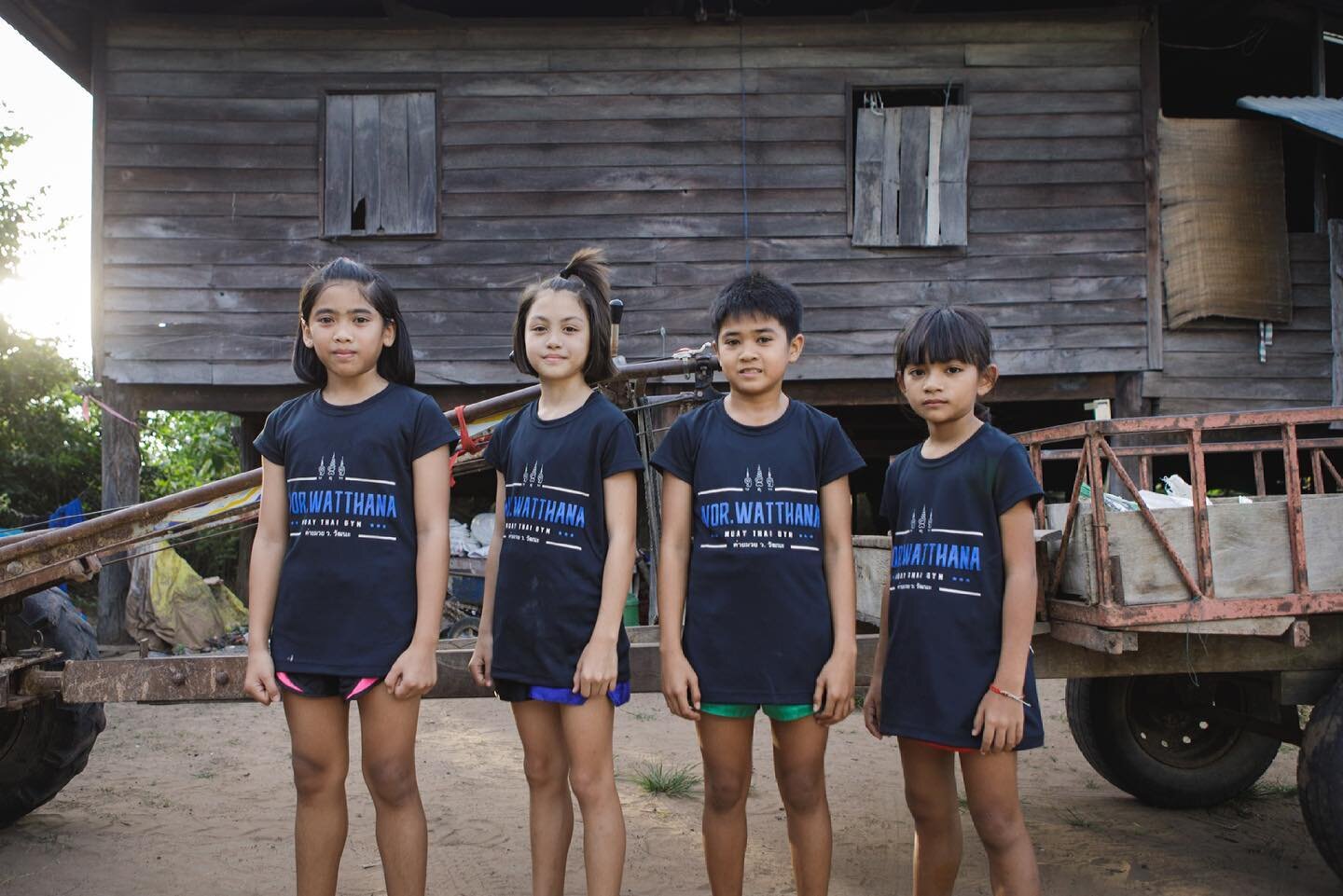 Four children in Wor. Watthana shirts standing in front of a traditional wooden house in the village.