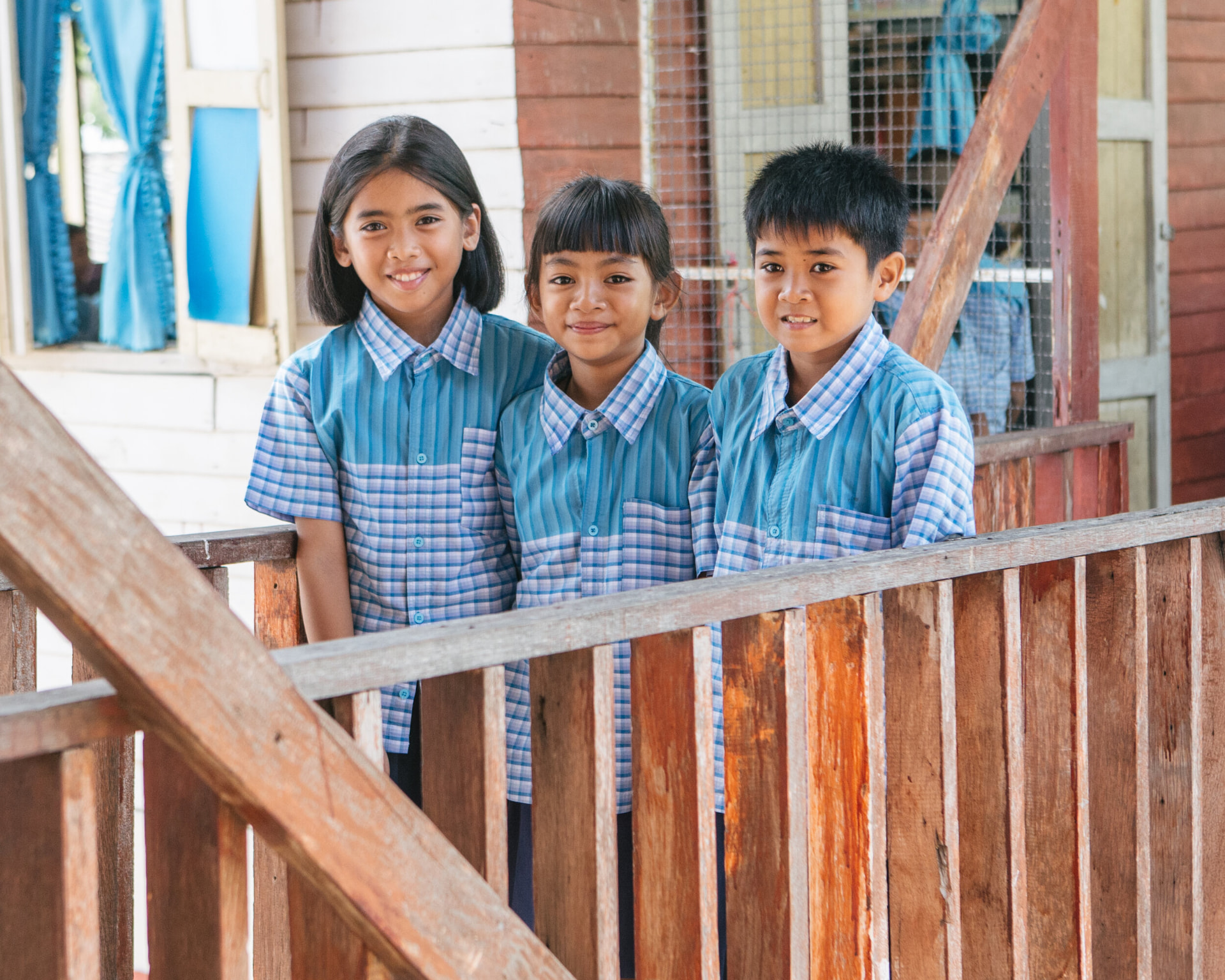 Three children in school uniforms smiling together on the wooden steps of their school.